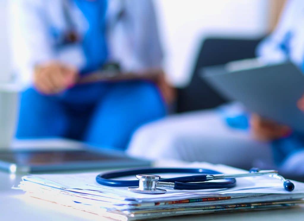 A stack of reports with a stethoscope and blurred view of people talking