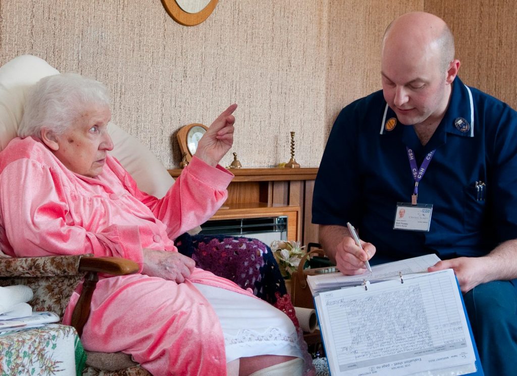 An elderly patient in pink with a male district nurse filling in report