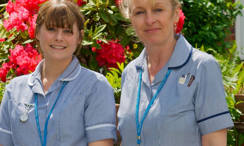 Two community nurses outside with flowers in hedge behind them in the summer