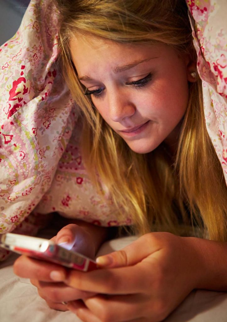 A sad looking teenage girl with blond hair texting using a mobile phone under covers