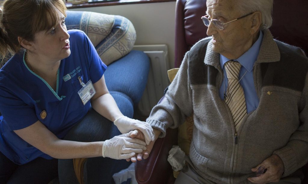 An elderly man with asthma talks with a district nurse as she reassures him and holds his hand