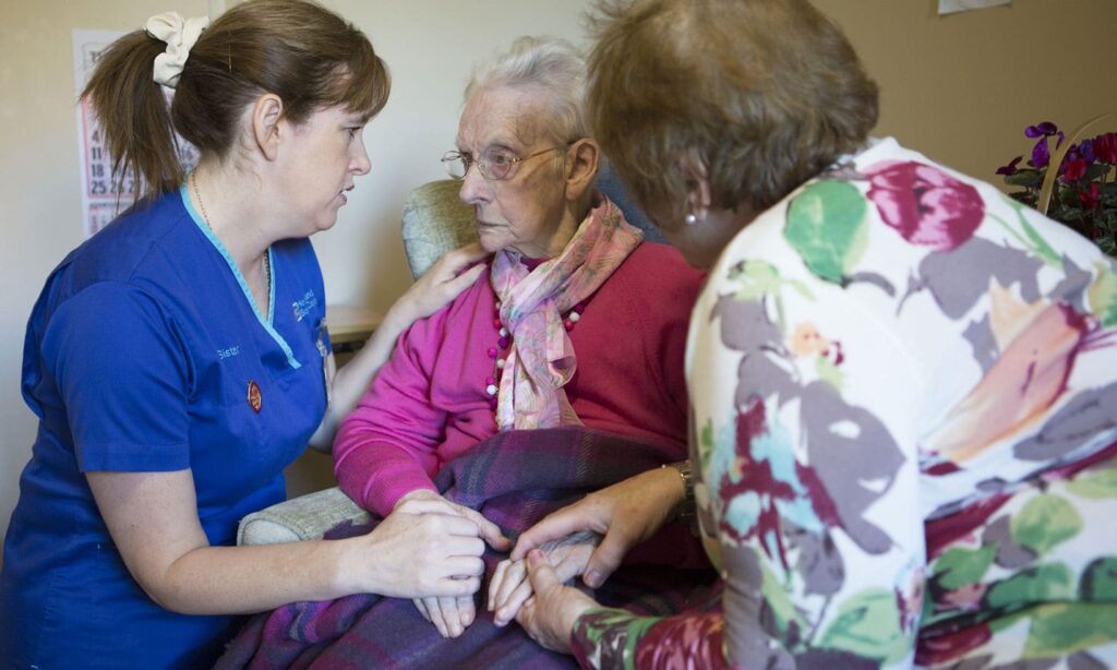 A nurse talking to an elderly woman with her carer in the foreground, holding her hands